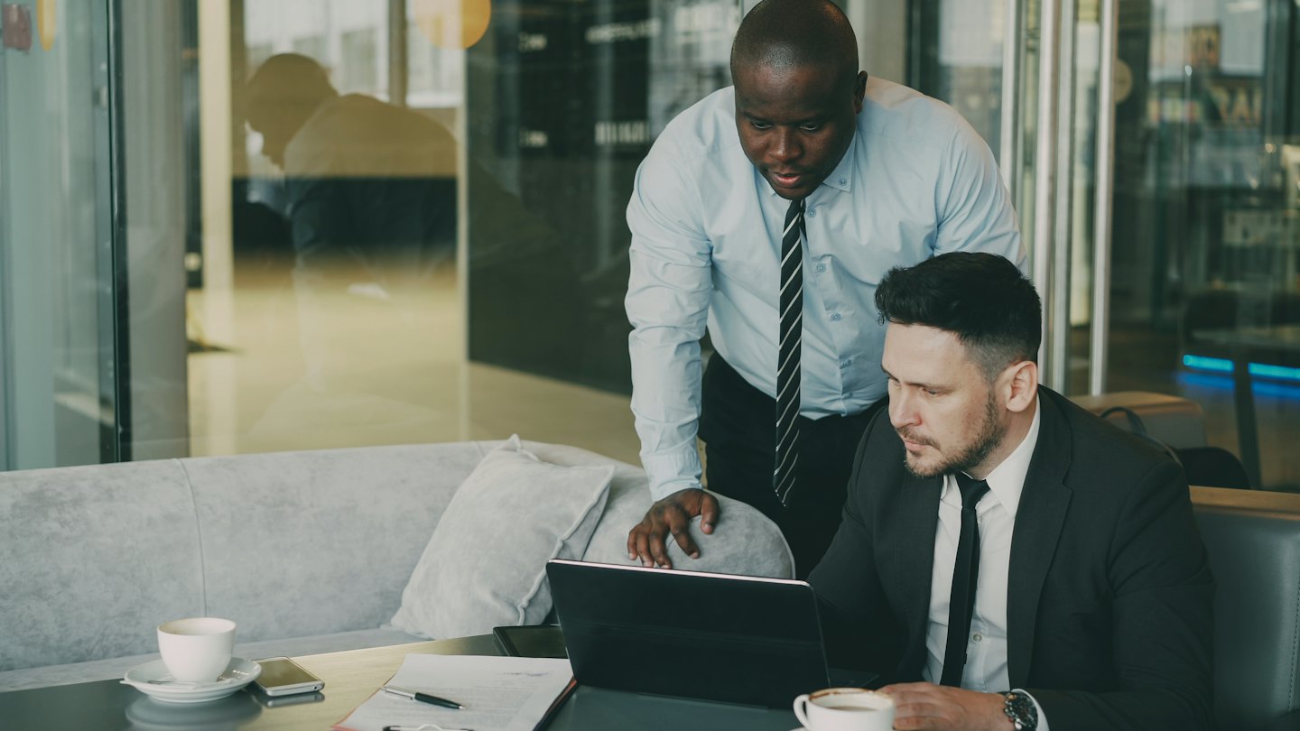 Two businessmen collaborate on a laptop at a table.